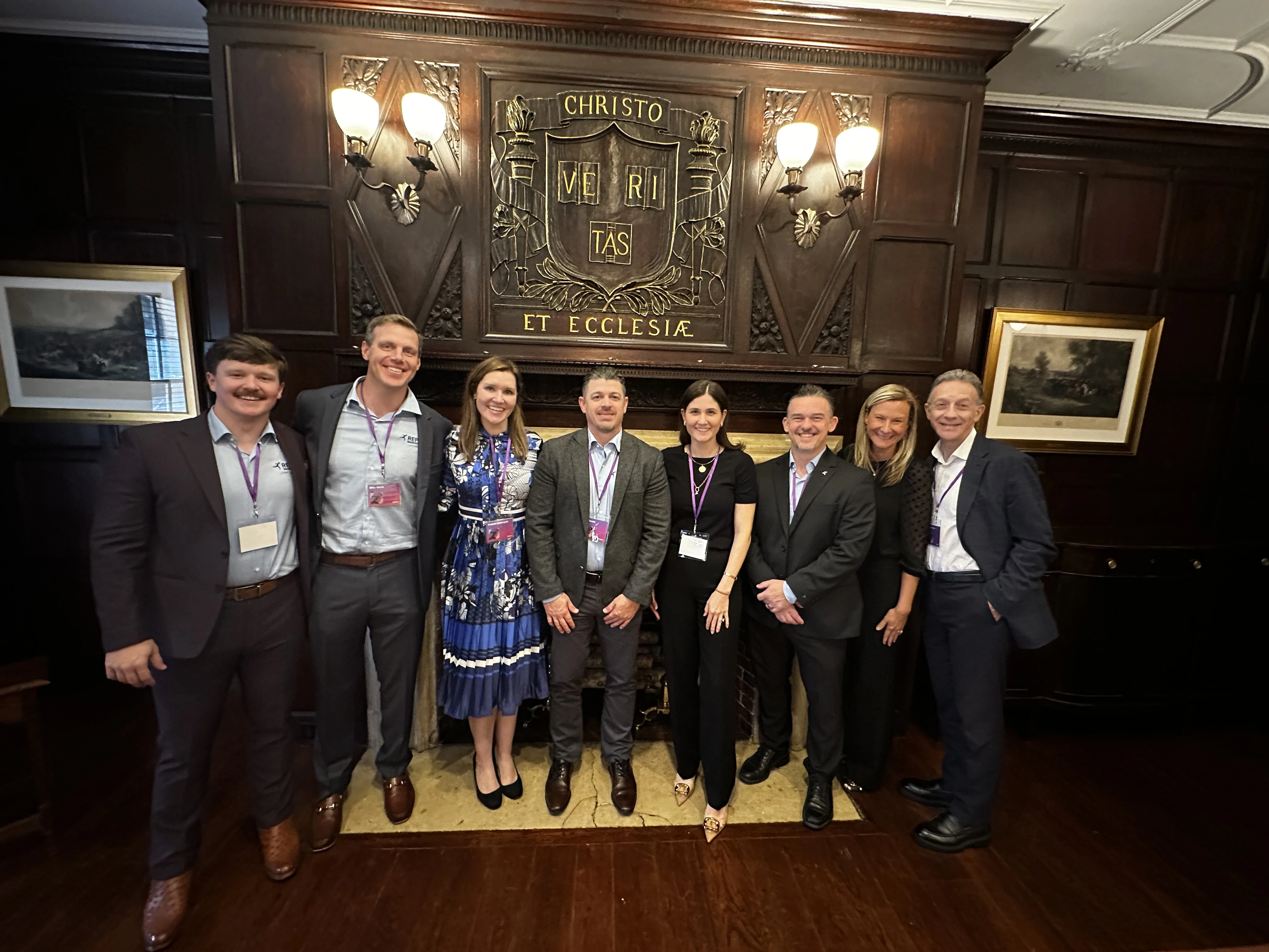 Sagepath Reply team dressed up and smiling in front of a wooden crest wall at Harvard Club during Opticon 2025.