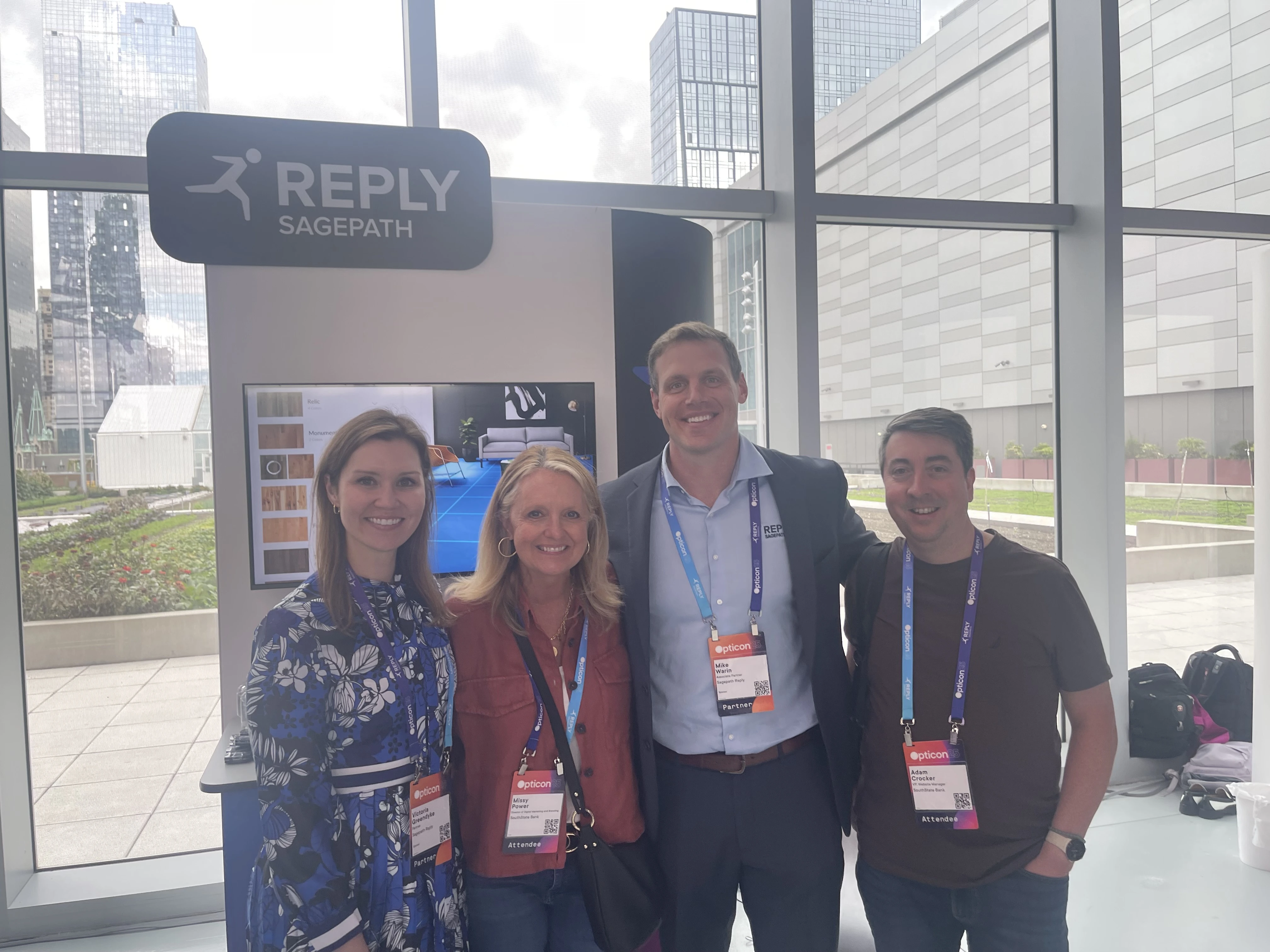 Sagepath Reply team and client posing by their display booth at Opticon 2025 with city buildings in the background.